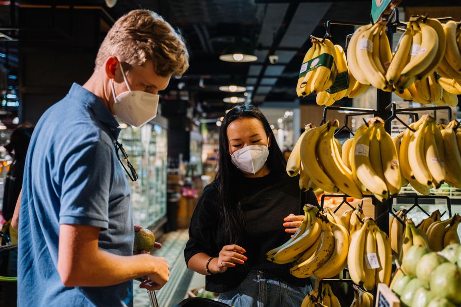Pessoas usando máscaras ao escolherem frutas em uma feira ou supermercado, destacando a segurança alimentar e o consumo consciente.