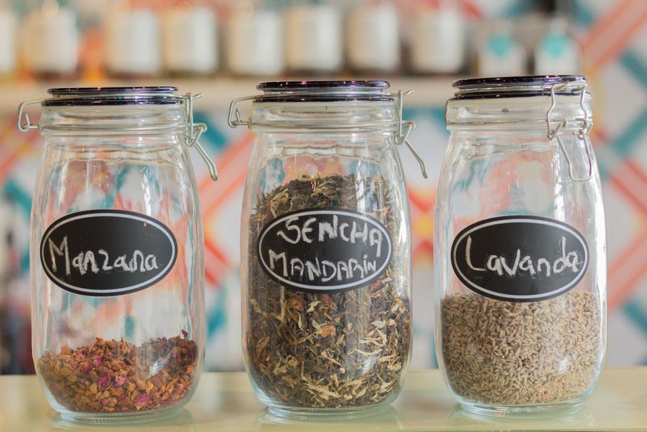 Três potes de vidro com diferentes ervas aromáticas rotulados à mão, usados para chá ou temperos, exibidos em uma loja ou dispensário de produtos naturais.