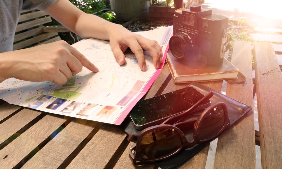 Pessoa planejando uma viagem ao ar livre em um ambiente de descanso, com câmera, óculos e celular ao lado, em um cenário ensolarado.