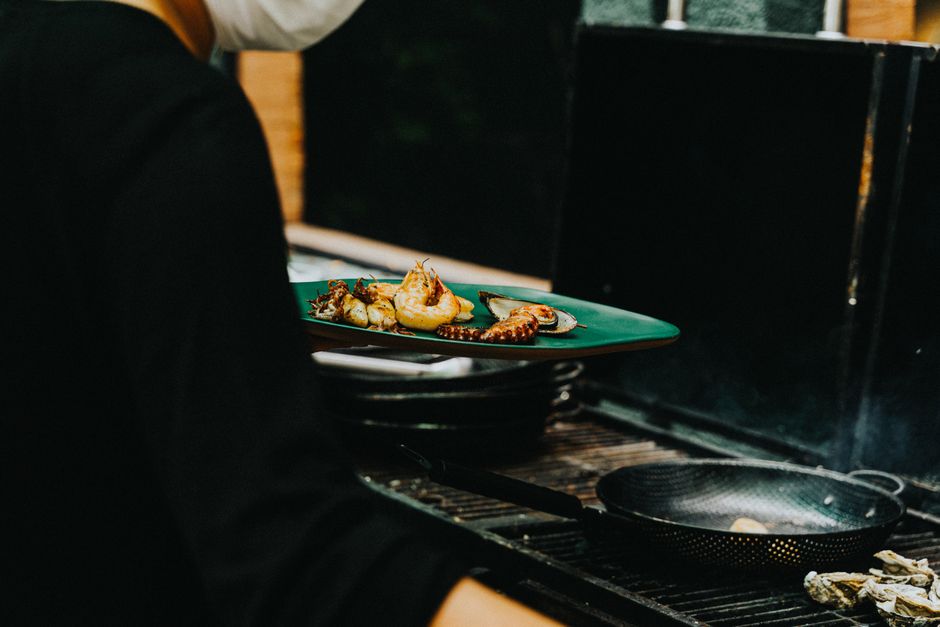Pessoa cozinhando frutos do mar em uma grelha ao ar livre, em um ambiente que lembra churrasqueira ou espaço externo para churrasco.