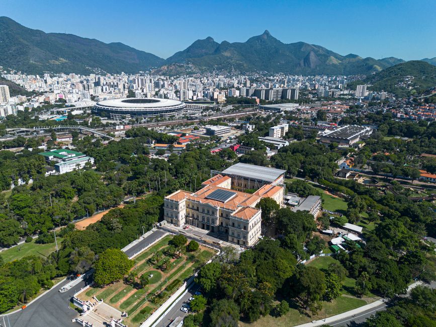 Vista aérea de um parque urbano com uma construção histórica ao centro, cercada por uma cidade moderna e montanhas ao fundo, representando o contraste entre história e urbanização na cidade do Rio de Janeiro.