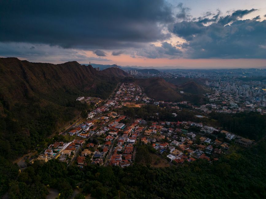 Vista aérea de uma cidade cercada por morros sob um céu com nuvens ao pôr do sol, evidenciando o contraste entre área urbana e natureza.