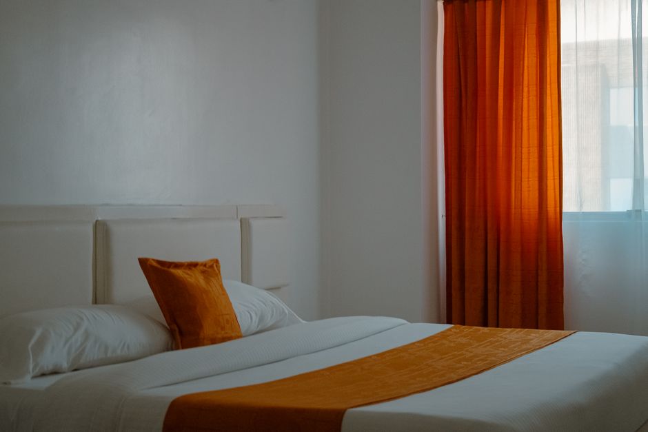 Quarto aconchegante com cama bem arrumada, almofada e cortinas em tons de laranja, transmitindo conforto e tranquilidade.