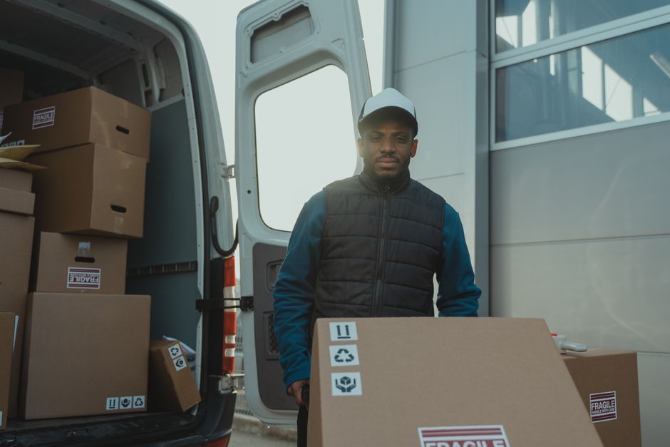 Entregador segurando uma caixa de transporte de encomendas ao lado de uma van de entrega, representando logística e entregas rápidas.