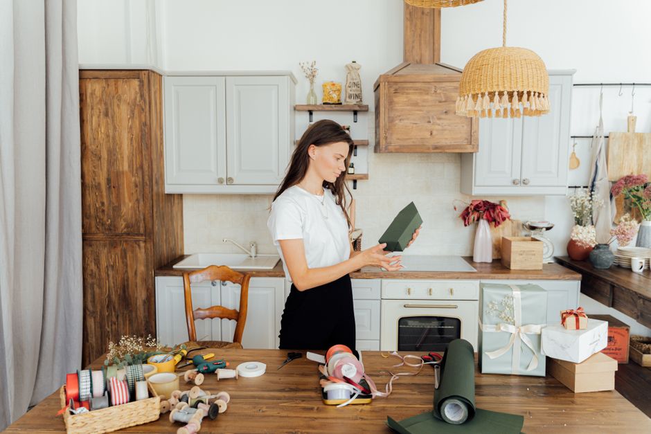 Mulher preparando uma embalagem de presente em uma cozinha decorada com elementos rústicos, em um ambiente acolhedor e bem iluminado.