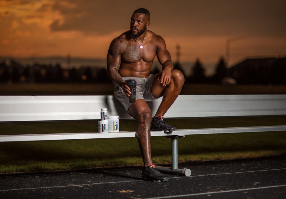 Homem musculoso, sem camisa, sentado em banco de parque ao pôr do sol, com suplementos esportivos ao lado, refletindo sobre treino e saúde.