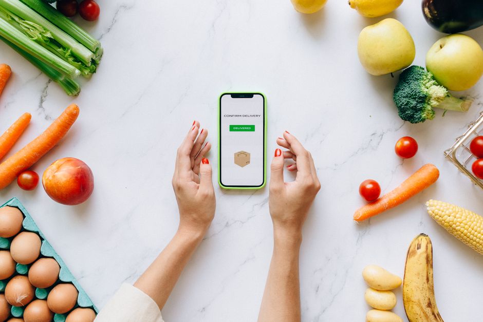 Pessoa usando aplicativo de delivery em smartphone rodeada de alimentos frescos sobre uma bancada de mármore.