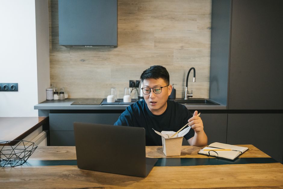 Jovem trabalhando de forma remota na cozinha, assistindo a conteúdo no computador enquanto faz sua refeição, refletindo o conceito de trabalho e lazer no home office.