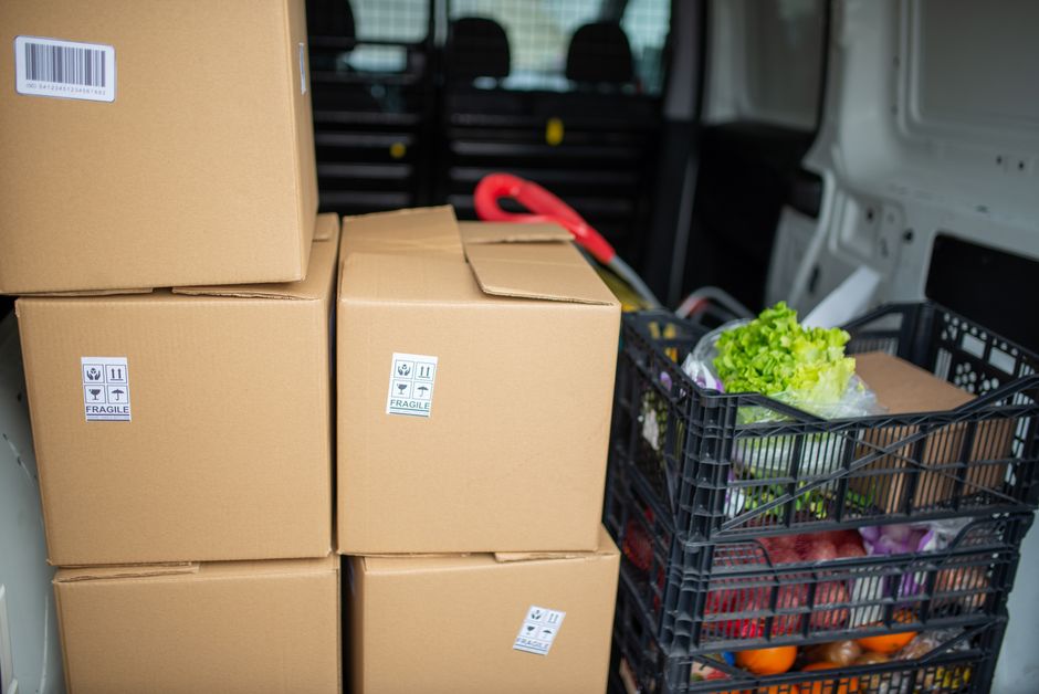 Imagem de caixas de papelão empilhadas ao lado de uma cesta repleta de hortifrúti dentro de uma van de entregas, representando logística de alimentos ou transportes de compras.