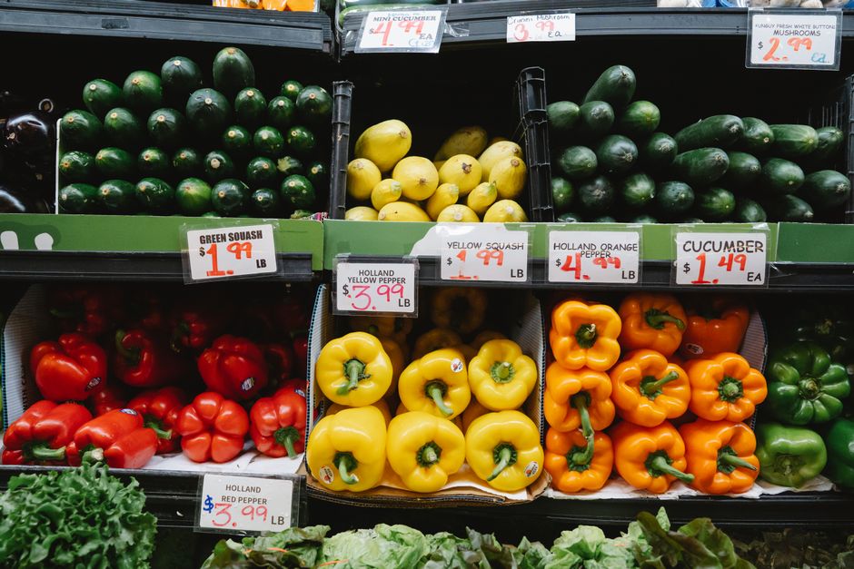 Estantes de hortifrúti exibindo uma variedade de pimentões, abóbora, pepino e outros vegetais em uma feira ou mercado.