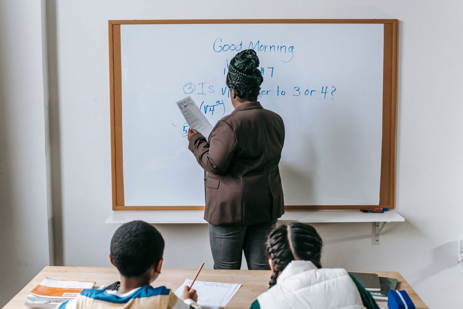 Professora explicando questões de matemática para alunos em sala de aula, com foco no ensino fundamental ou médio.