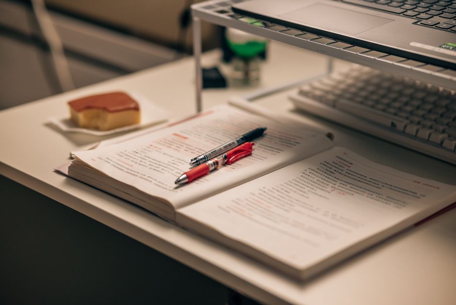 Imagem de um ambiente de trabalho com documentos, uma caneta, um computador portátil e um lanche ao lado, ideal para ilustrar o tema de estudos ou rotina no escritório.