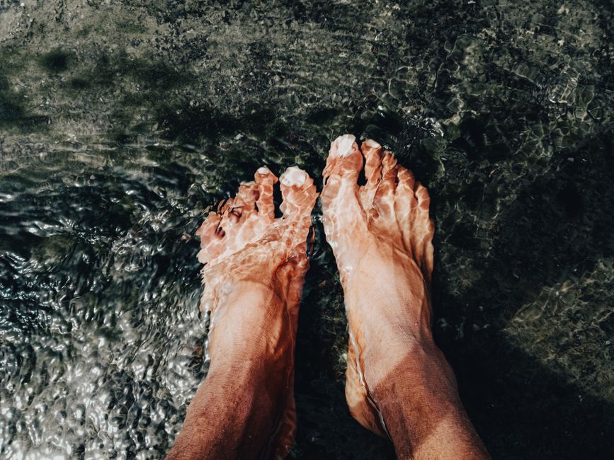 Pés mergulhados na água de uma praia de areia escura, transmitindo uma sensação de relaxamento e conexão com a natureza.