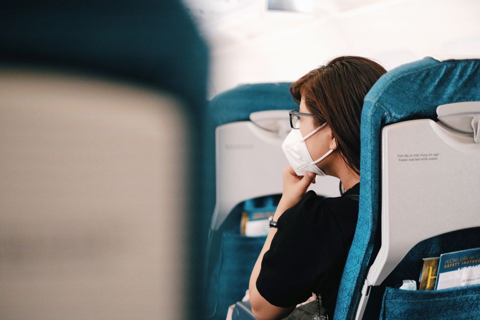 Mulher de máscara facial, usando óculos, sentada em assento de avião, refletindo ou descansando durante voo, representando viagem ou deslocamento.