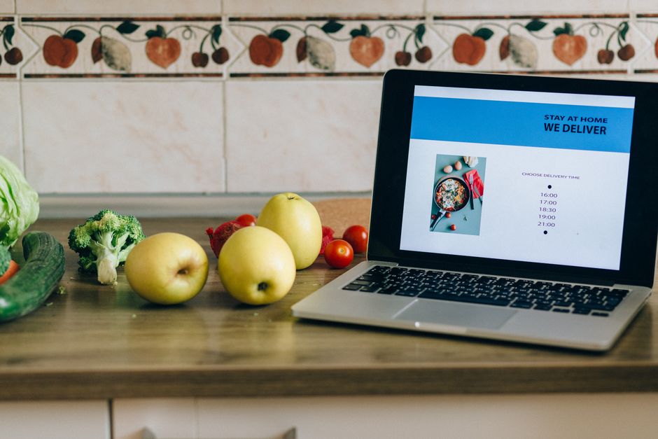 Imagem de uma cozinha com vegetais ao lado de um laptop exibindo uma tela de delivery de comida, sugerindo tema de alimentação saudável e compras online.