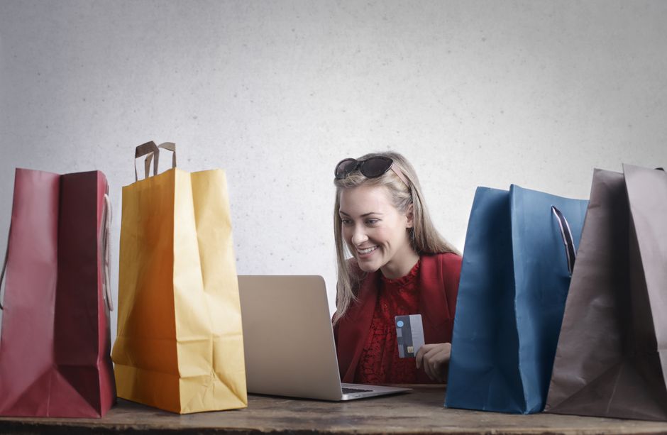 Mulher realiza compras online com várias sacolas de lojas ao redor, representando varejo e e-commerce.