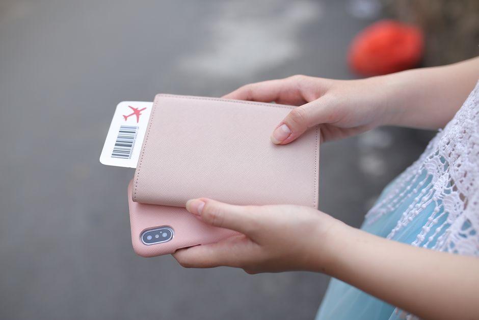 Pessoa segurando um smartphone rosa com capa de couro e um cartão de embarque de avião, pronta para viajar, em ambiente ao ar livre.