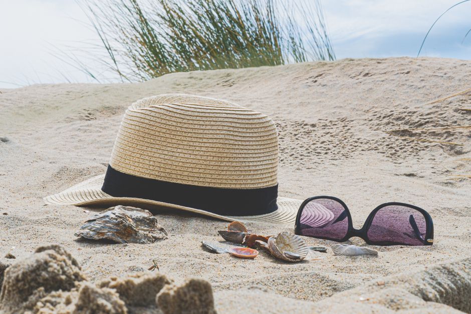 Imagem de chapéu de praia e óculos de sol na areia de uma praia, remetendo a um tema de férias e lazer ao ar livre.