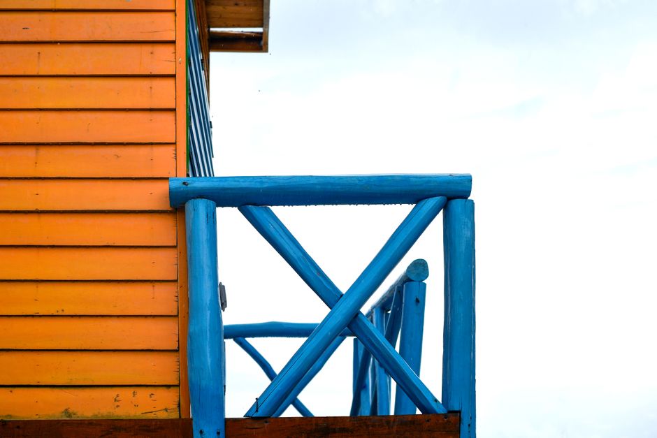 Foto de uma varanda de cabana de madeira com estrutura de suporte azul, destacando o contraste de cores entre a parede laranja e a estrutura azul sob céu claro, ideal para ilustrar temas de lazer ao ar livre ou vida na natureza.