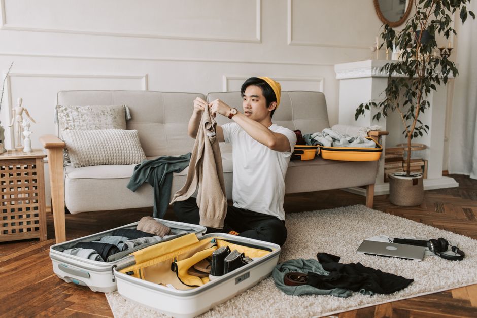 Jovem organizando roupas e objetos pessoais para uma viagem, preparando a mala em um ambiente de sala de estar.