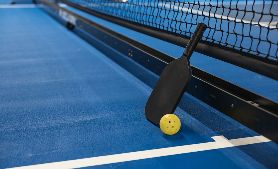 Imagem de uma quadra de tênis de mesa com uma raquete e uma bola amarela ao lado da rede, representando o tema de esportes e lazer.