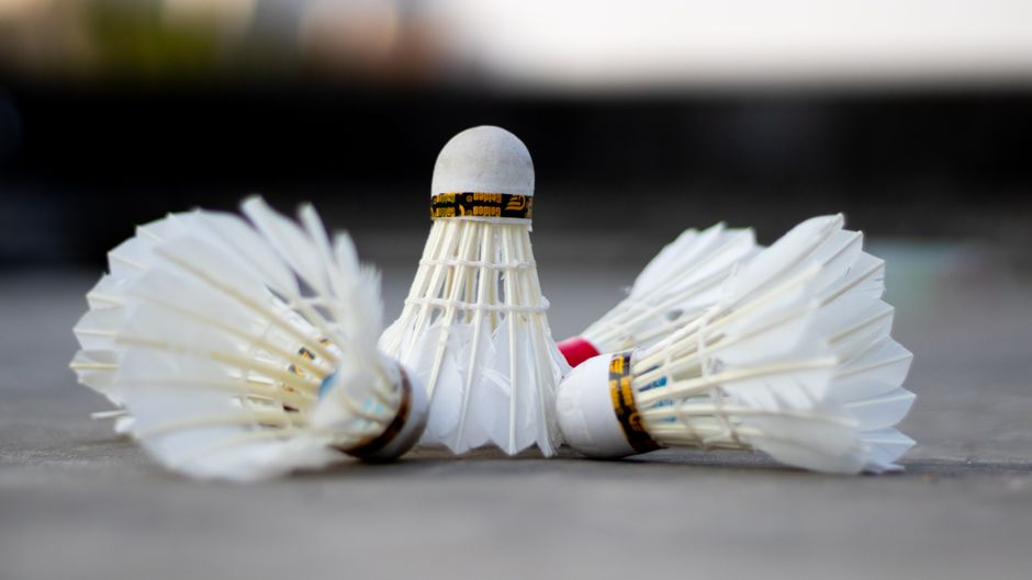 Imagem com três raquetes de badminton ao redor de uma bolinha de plástico, simbolizando competição ou esporte ao ar livre.