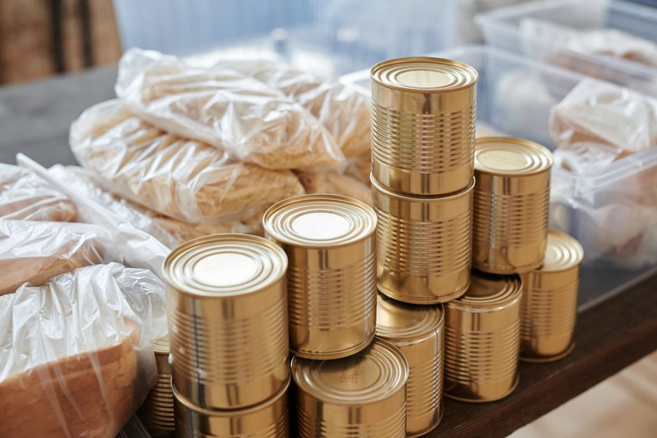 Imagem de várias latas de alimentos empilhadas ao lado de alimentos embalados, sugerindo estoque ou preparação para distribuição de alimentos.