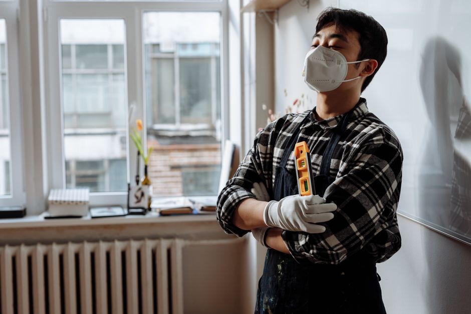 Jovem profissional de manutenção ou reparo pensando enquanto segura uma ferramenta em um ambiente de escritório ou sala de aula, com uso de máscara e luvas.