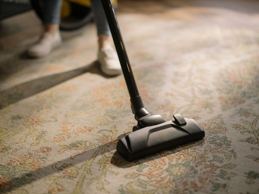 Pessoa usando aspirador de pó em uma fibra de tapete de aparência floral, demonstrando limpeza e manutenção de ambientes internos.