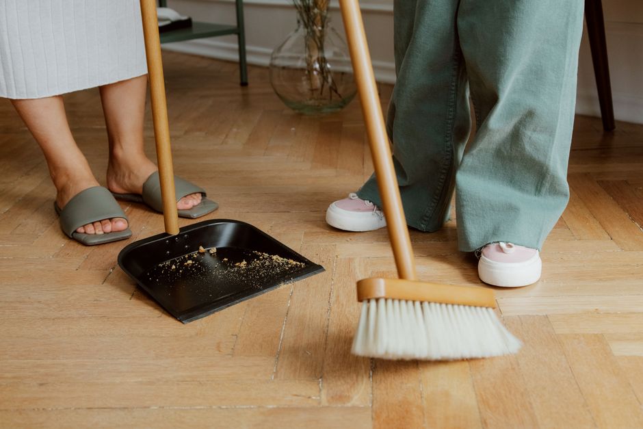 Pessoas limpando o chão de madeira com vassoura e pá de lixo em ambiente interno.