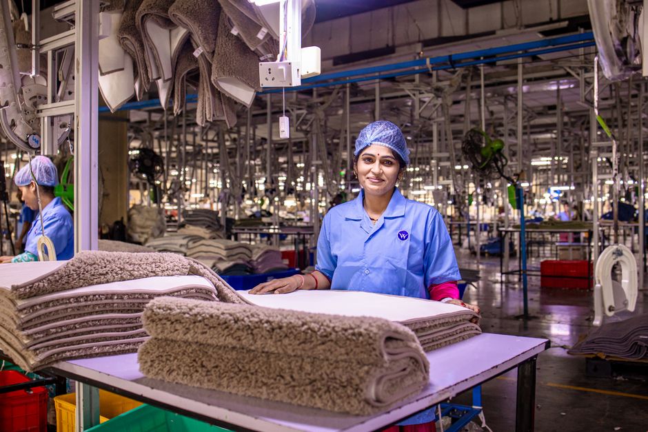 Funcionária em fábrica de tecidos durante inspeção de produção, destacando o ambiente industrial e trabalhos manuais relacionados à confecção de tapetes ou mantas.