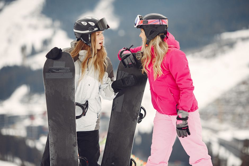Duas mulheres conversando e se apoiando durante uma atividade de snowboard em uma paisagem de inverno com montanhas ao fundo.