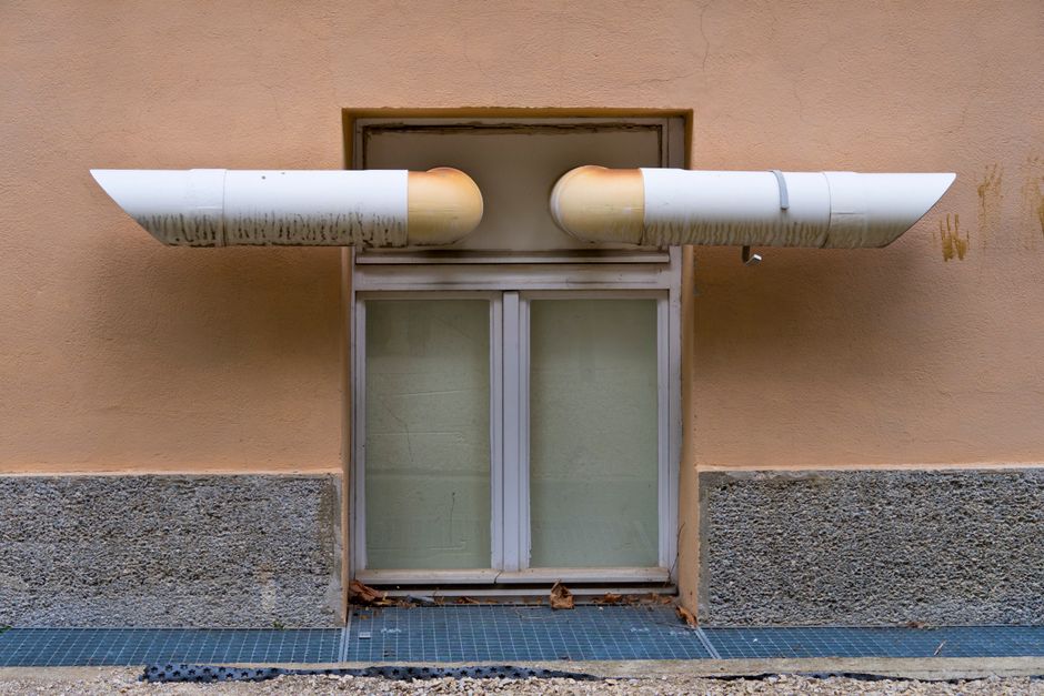 Imagem de um sistema de exaustão de ar externo, com duas tubulações de ventilação que saem da parede de um edifício, sugerindo infraestrutura de ventilação ou climatização.