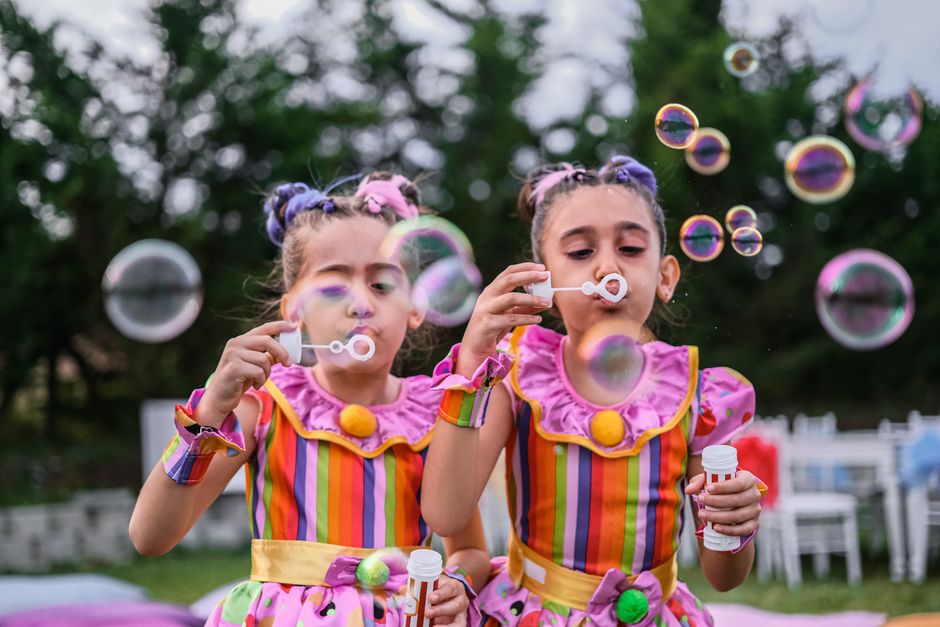 Duas meninas brincando de fazer bolhas de sabão em um evento ao ar livre, vestidas com fantasias coloridas e alegres.
