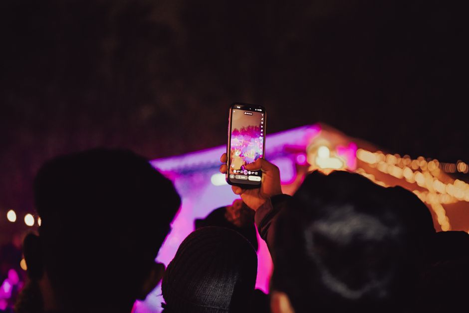 Pessoa registrando um show ao vivo com o celular durante a noite, com luzes coloridas e público ao fundo.
