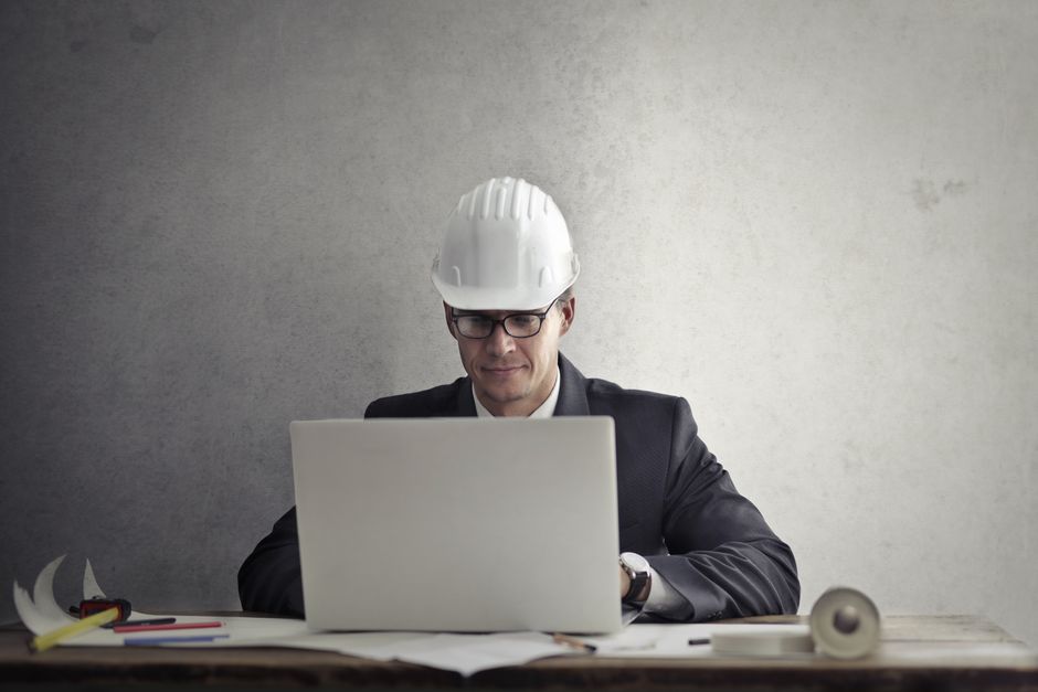 Homem de terno com óculos e capacete de segurança sentado em frente ao computador, simbolizando planejamento e gestão de projetos de construção ou engenharia.