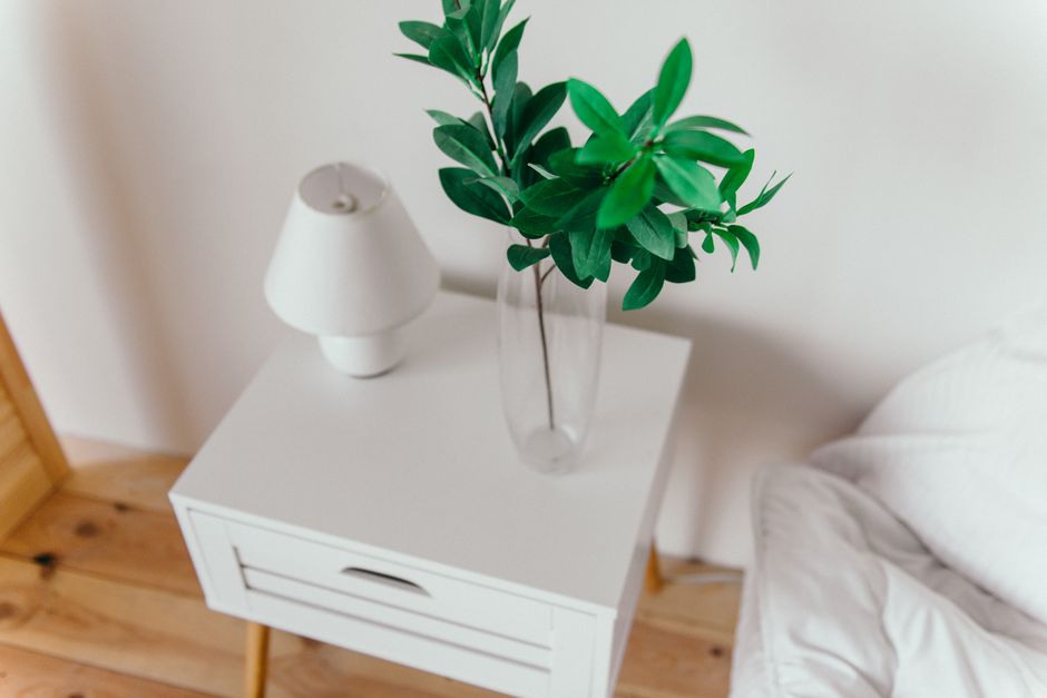 Imagem de uma cômoda branca ao lado de uma cama com uma planta verde em um vaso de vidro e uma luminária, em um ambiente de quarto minimalista.