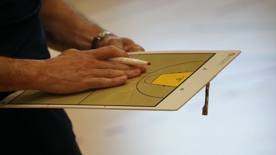 Pessoa usando uma prancheta para traçar estratégias táticas em uma quadra de basquete, destacando planejamento esportivo e análise de jogo.