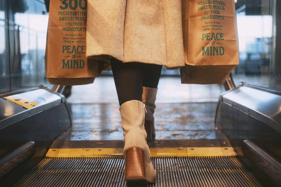 Mulher com botas e casaco carregando sacolas de supermercado ao sair de uma loja, simbolizando compras e consumo consciente.