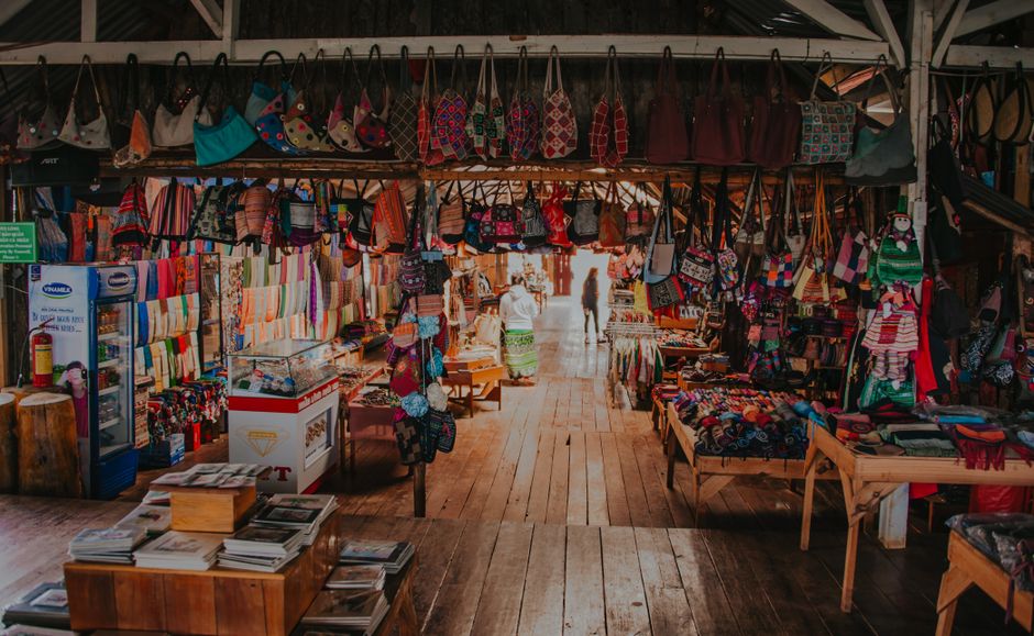 Imagem de uma feira de artesanato ao ar livre exibindo bolsas, roupas e acessórios coloridos, com pessoas ao fundo aproveitando o ambiente.