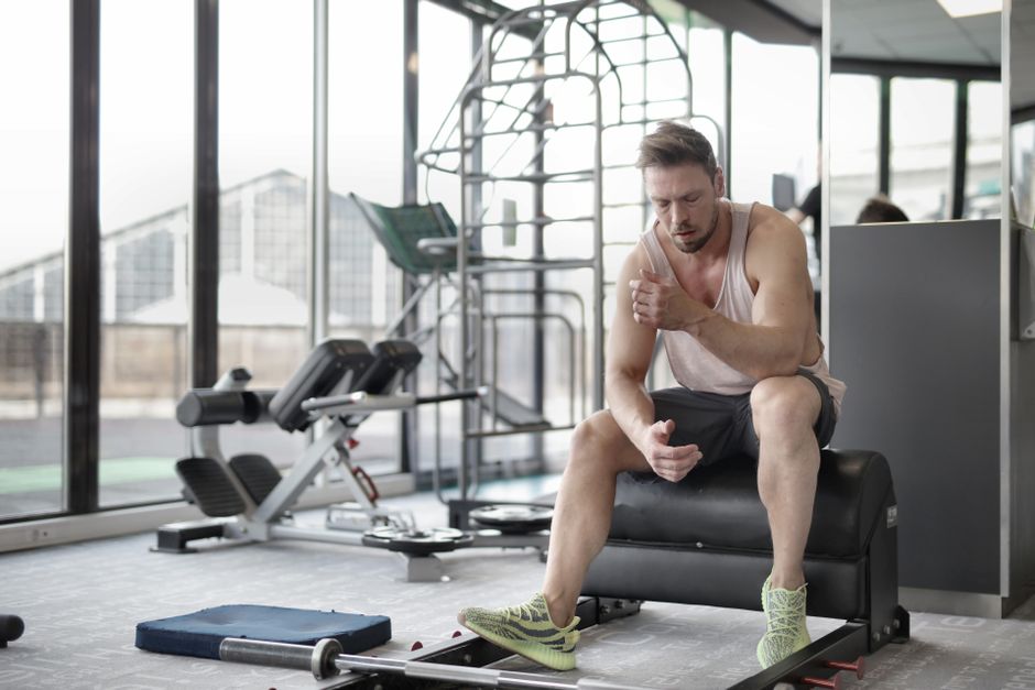 Homem descansando após exercício intenso em academia luminosa com equipamentos de treino ao fundo.
