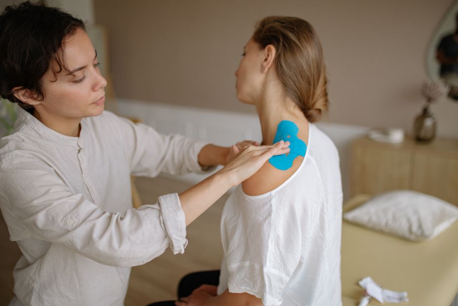 Mulher aplicando uma fita de colágeno na região do ombro de outra mulher, em ambiente interno, sugerindo um procedimento de cuidados estéticos ou tratamentos de saúde.