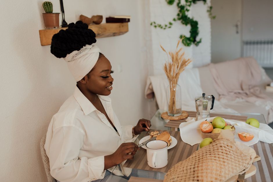 Mulher sorridente tomando café da manhã em ambiente acolhedor, com frutas e elementos decorativos naturais ao redor.