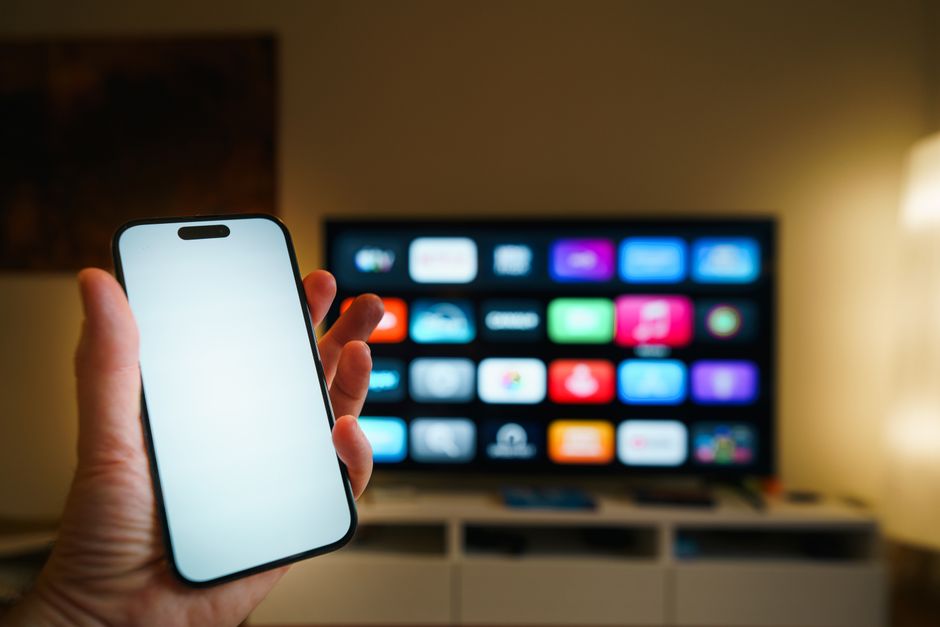 Imagem de uma pessoa segurando um smartphone com uma televisão ao fundo exibindo diversos ícones de aplicativos, ideal para artigos sobre inovação tecnológica e entretenimento digital.