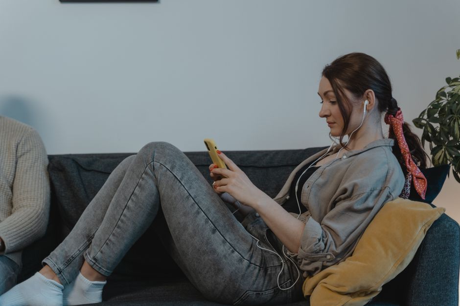 Mulher usando fones de ouvido e celular, relaxando em uma sala de espera ou ambiente informal, enquanto utiliza o celular.