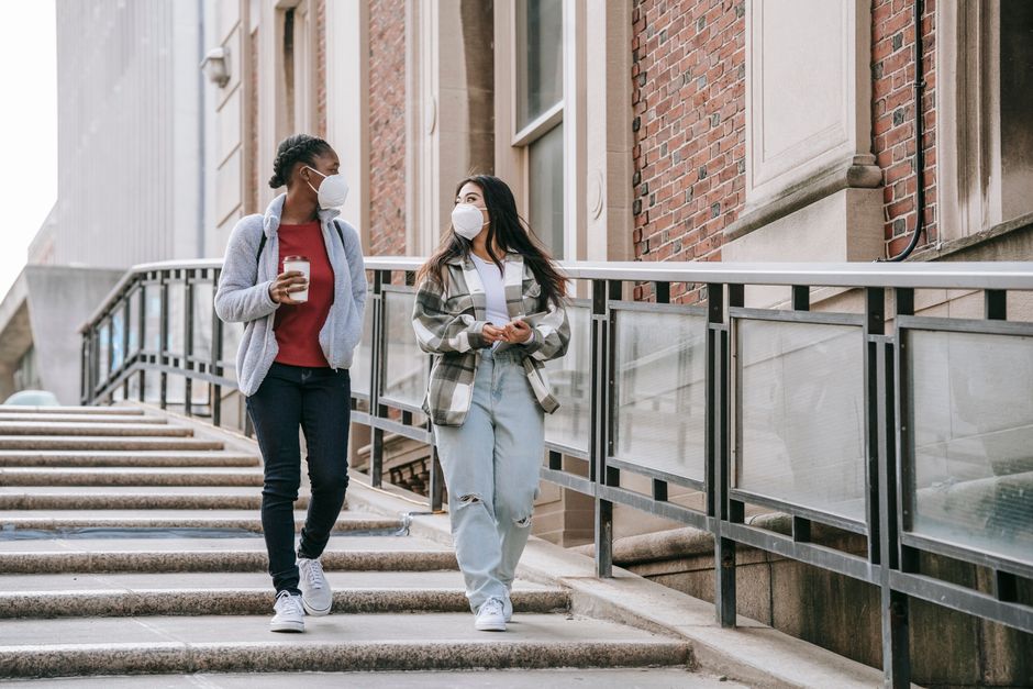 Duas mulheres caminhando e conversando ao ar livre na cidade, ambas usando máscaras faciais, simbolizando a retomada de atividades sociais com atenção às medidas de proteção contra a COVID-19.