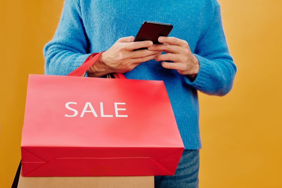 Pessoa realizando compras online para aproveitar uma liquidação, sustentando sacola de loja com destaque para a palavra SALE.