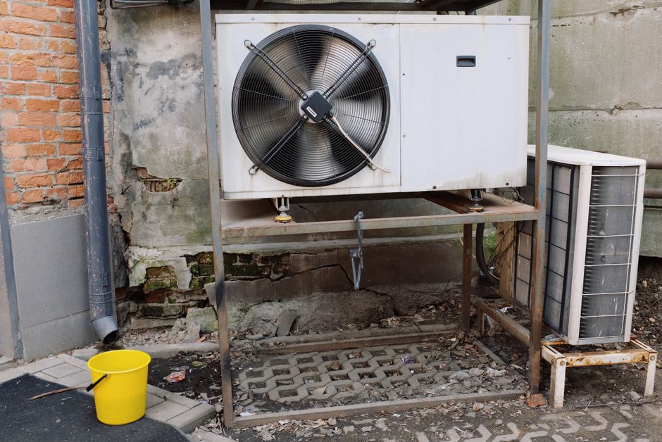 Imagem de uma unidade de ar condicionado instalada ao ar livre em uma parede de alvenaria com estrutura metálica e piso de concreto, indicando manutenção ou instalação de sistemas de resfriamento.