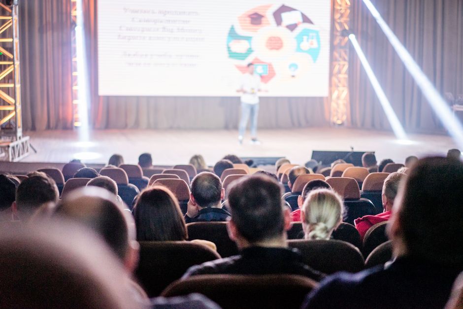 Imagem de uma apresentação ou palestra em um auditório, com foco na plateia assistindo a uma conferência ou evento de negócios.