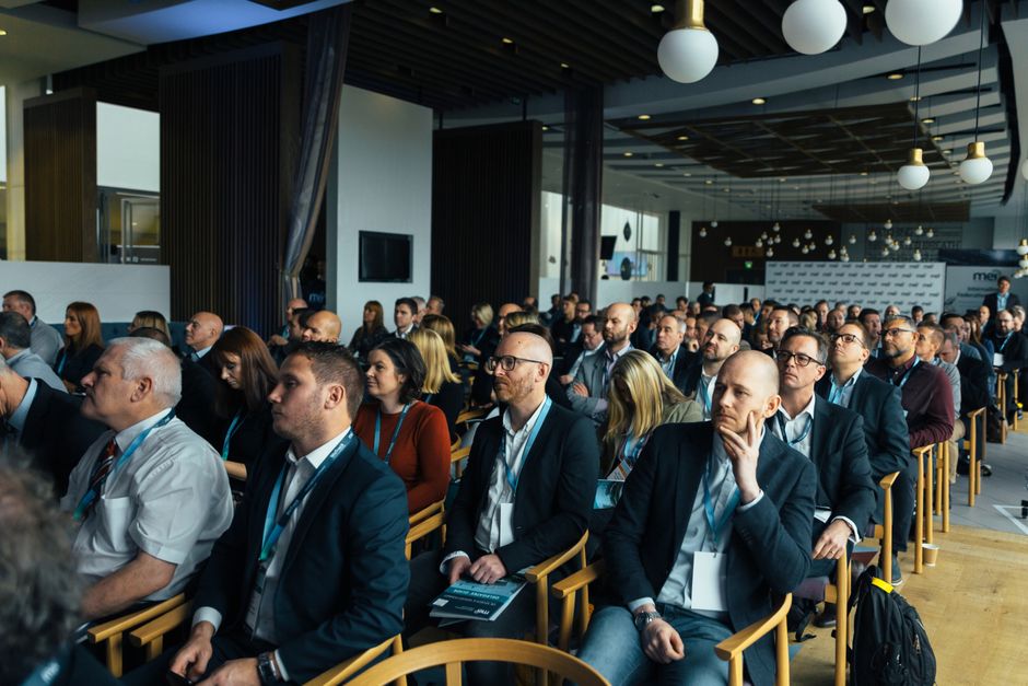 Imagem de um público de profissionais atentos durante uma conferência ou palestra empresarial em um ambiente moderno e bem iluminado.